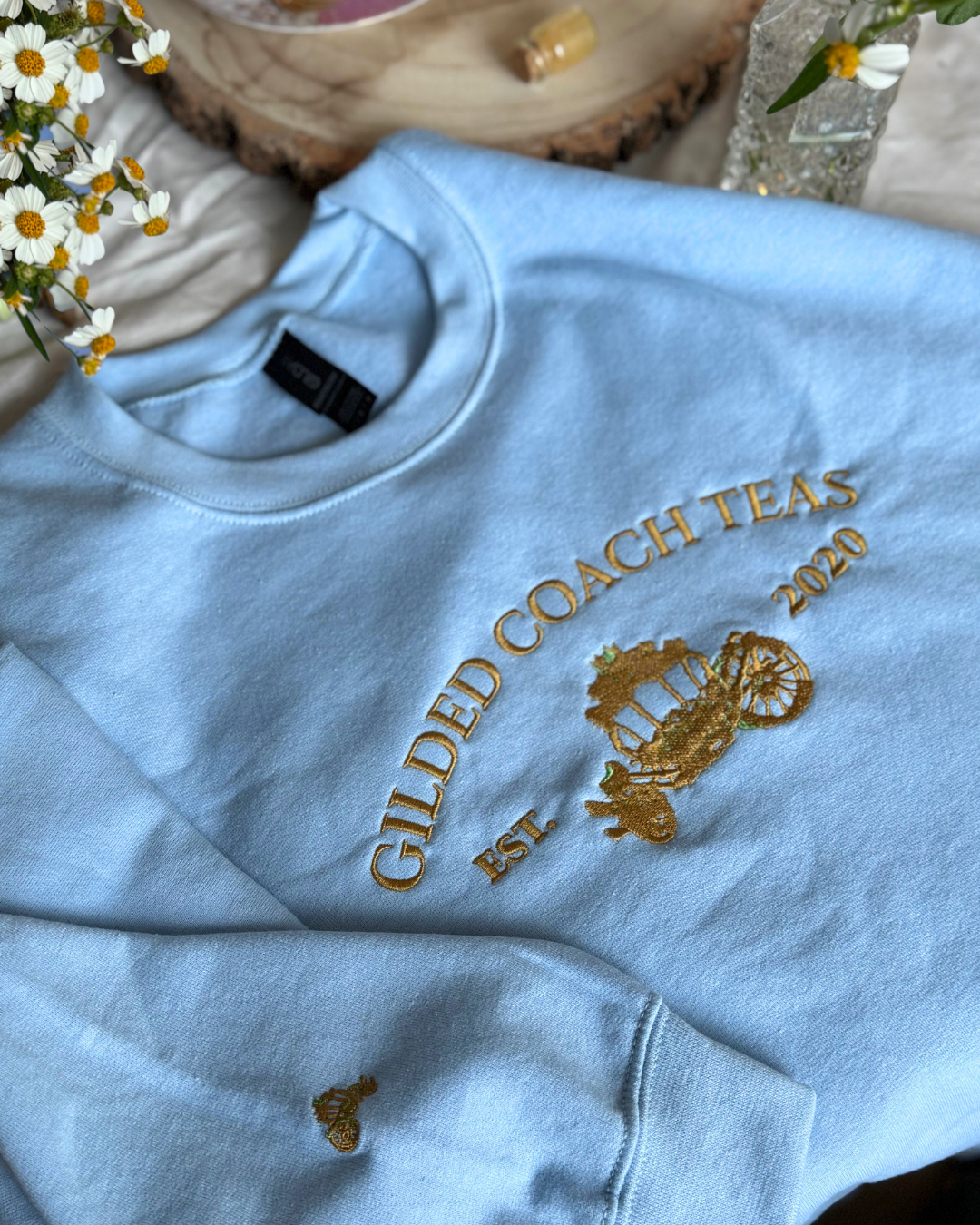 Light blue sweatshirt with gold text and logo on a surface with flowers in the background